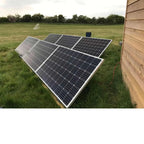 Ground-mounted solar panels on aluminum triangle frames in a grassy field next to a wooden building.