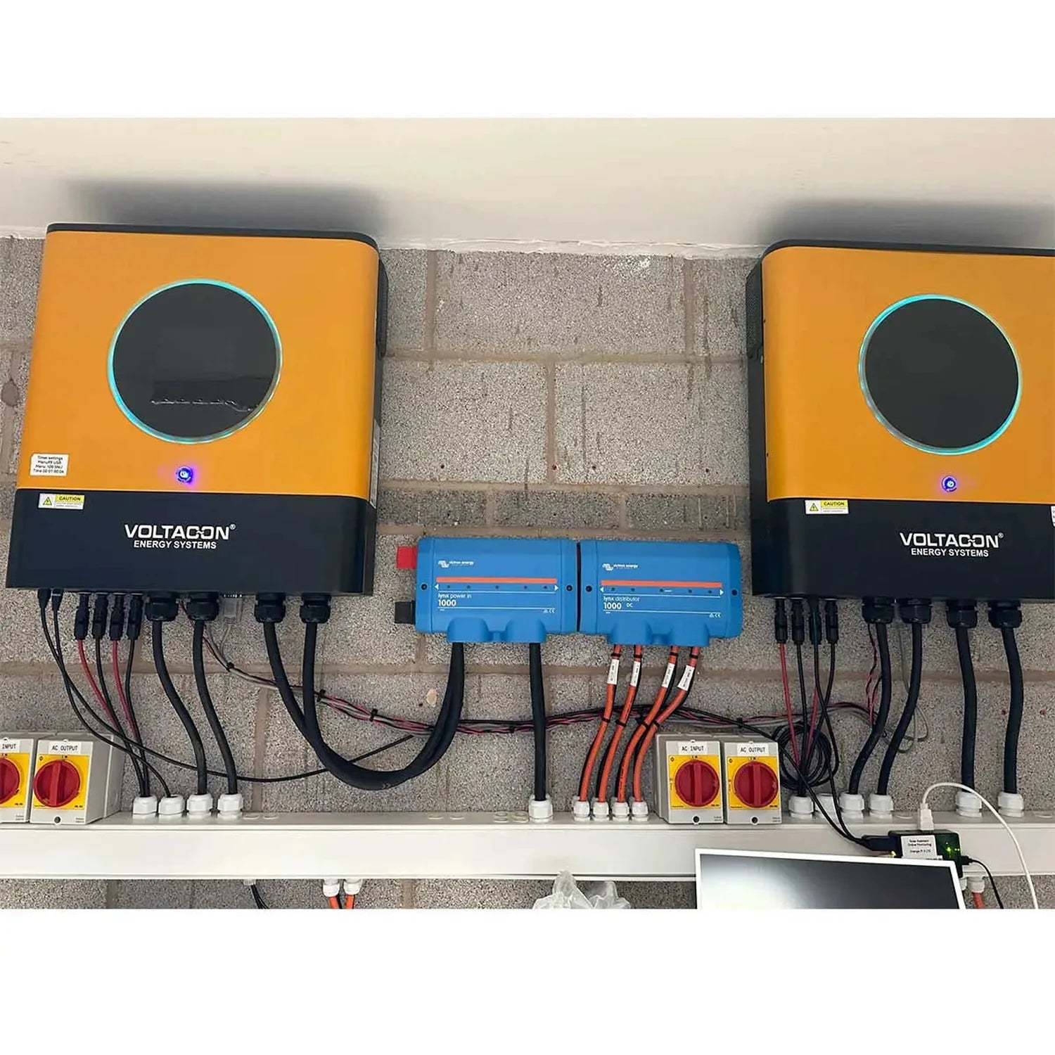Installation shot of two orange and black Voltacon Energy Systems inverters mounted on brick wall with blue charge controller