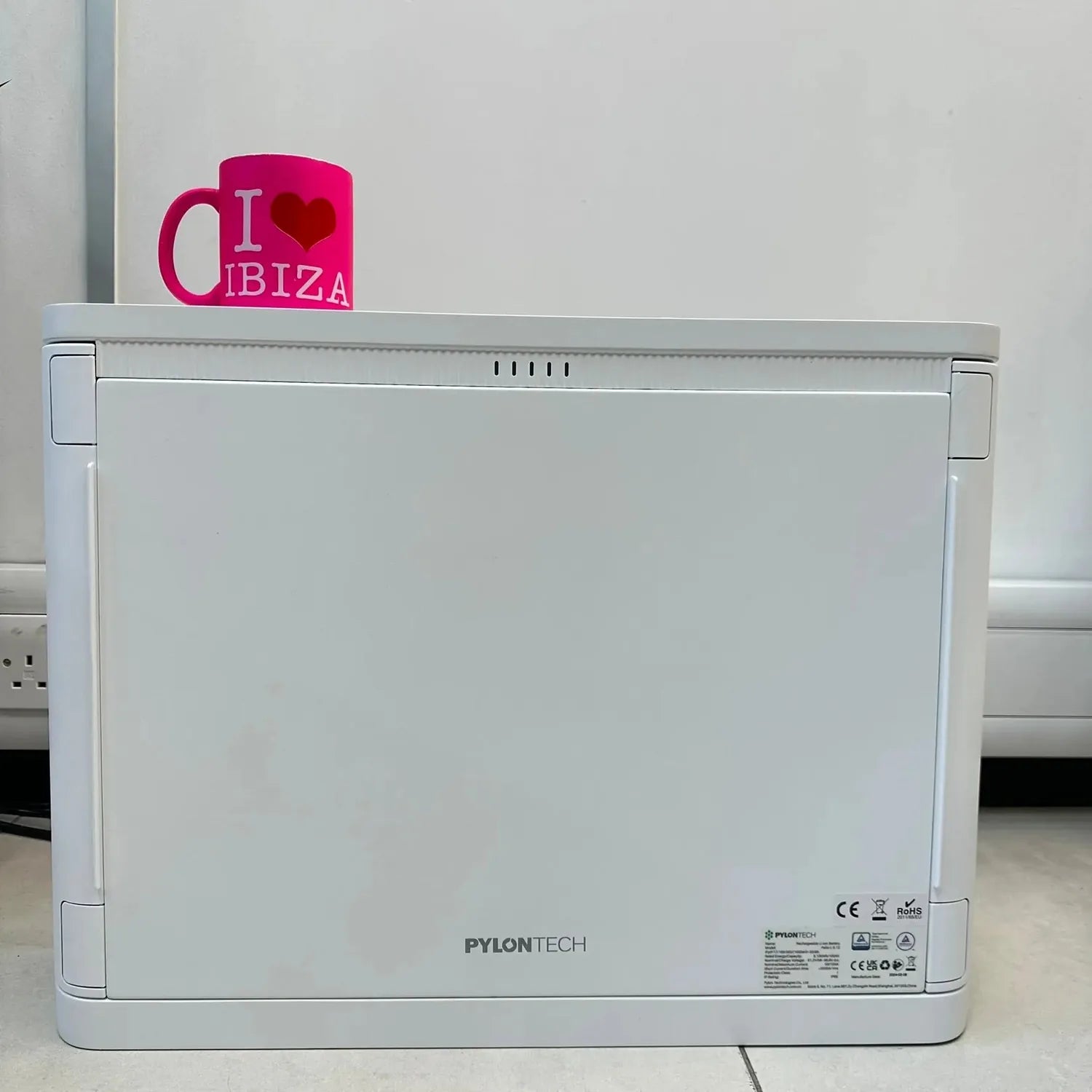 White Pylontech Pelio-L battery unit with pink "I love Ibiza" mug on top, showing rear panel with CE and RoHS certification l