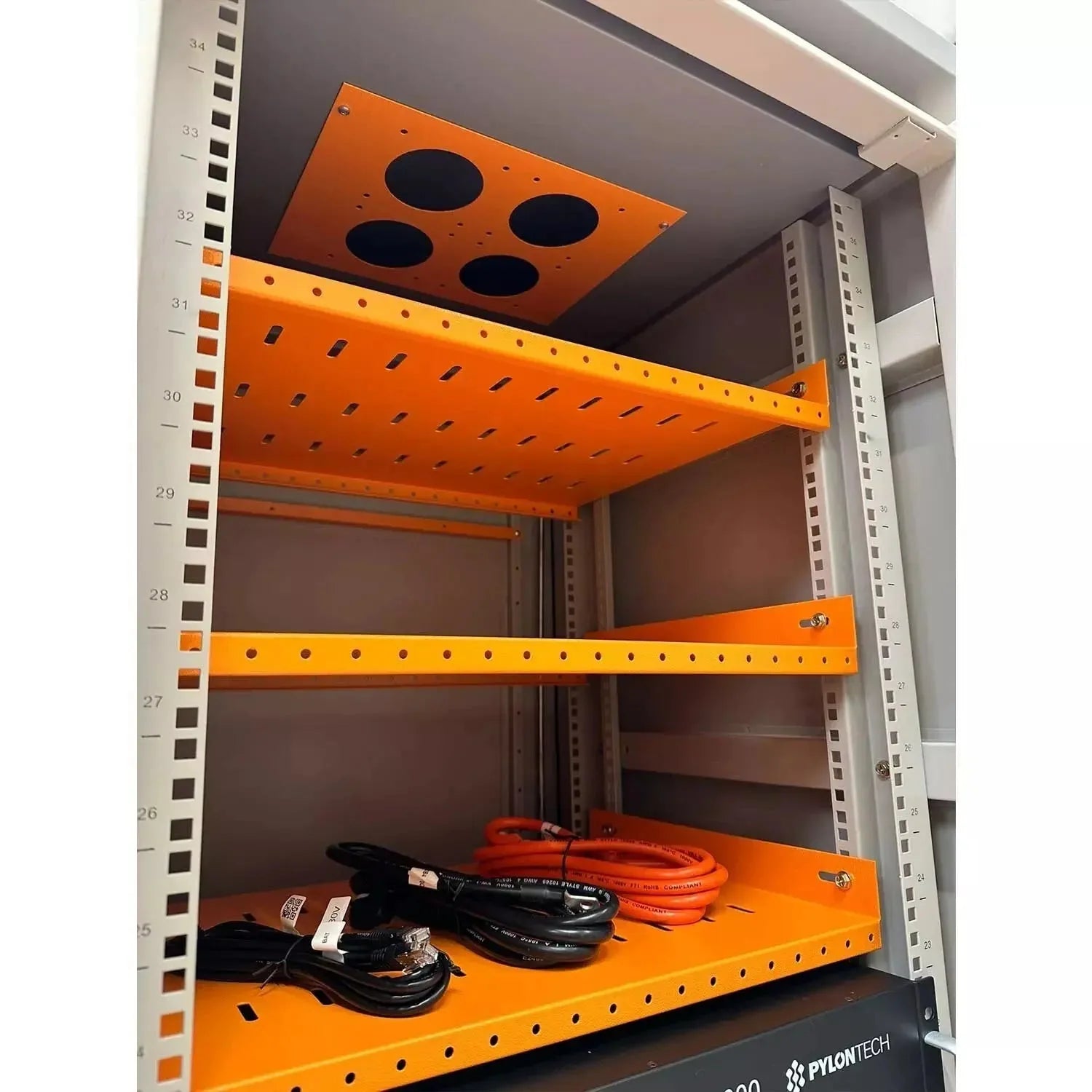 Orange metal battery enclosure mounted in white industrial rack with ventilation holes and cable management, containing black