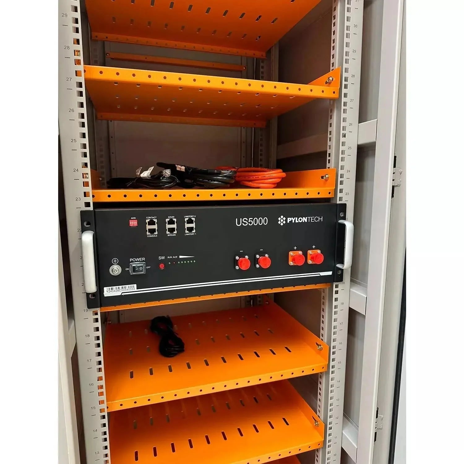 Pylon Tech US5000 battery enclosure mounted in white server rack with orange shelving and cables stored above.