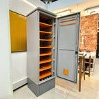Gray metal battery enclosure with orange shelving and open door, displayed in showroom with brick wall background.