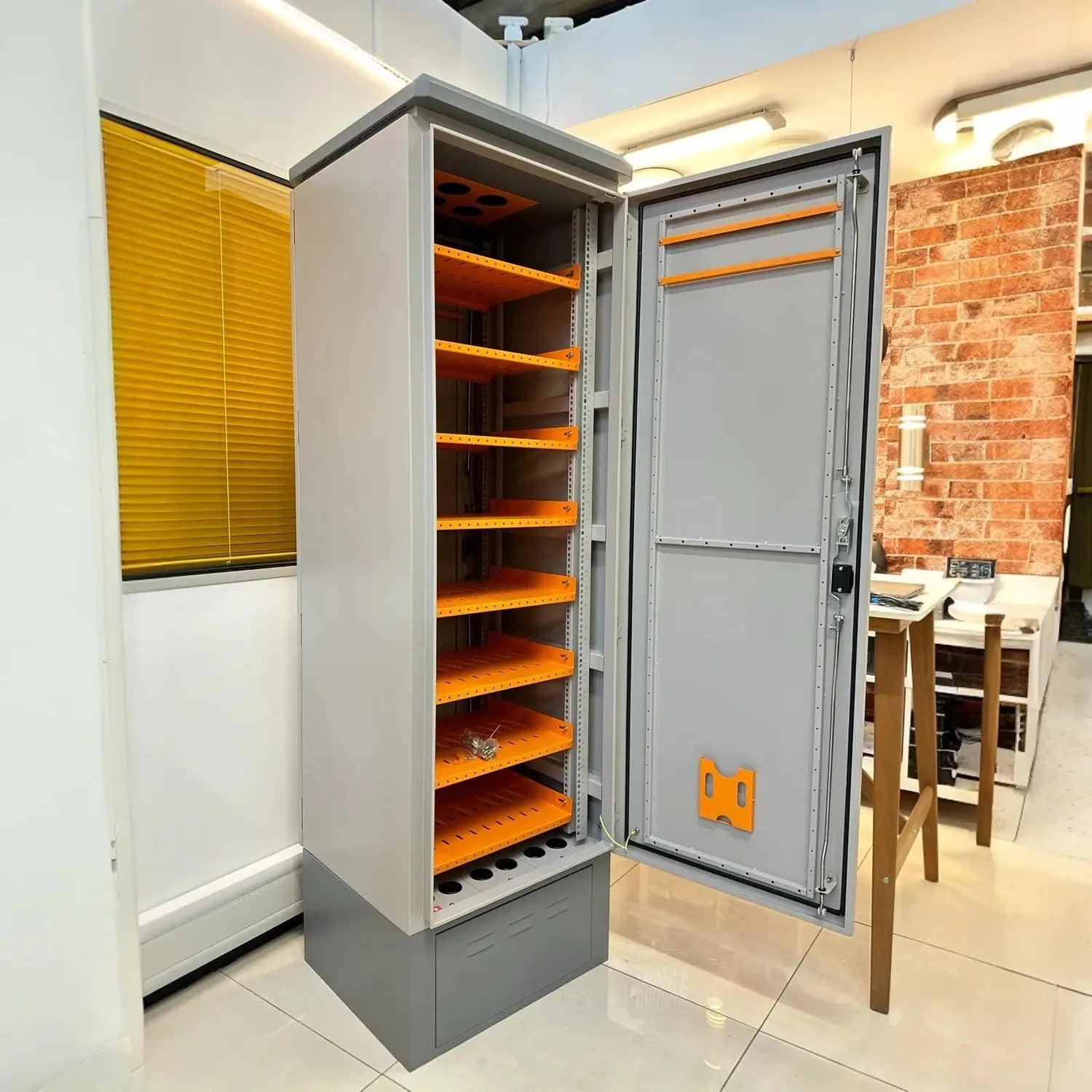 Gray metal battery enclosure with orange shelving and open door, displayed in showroom with brick wall background.