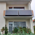 Two solar panels mounted on a modern white building's balcony railing, angled toward the sun with adjustment brackets visible