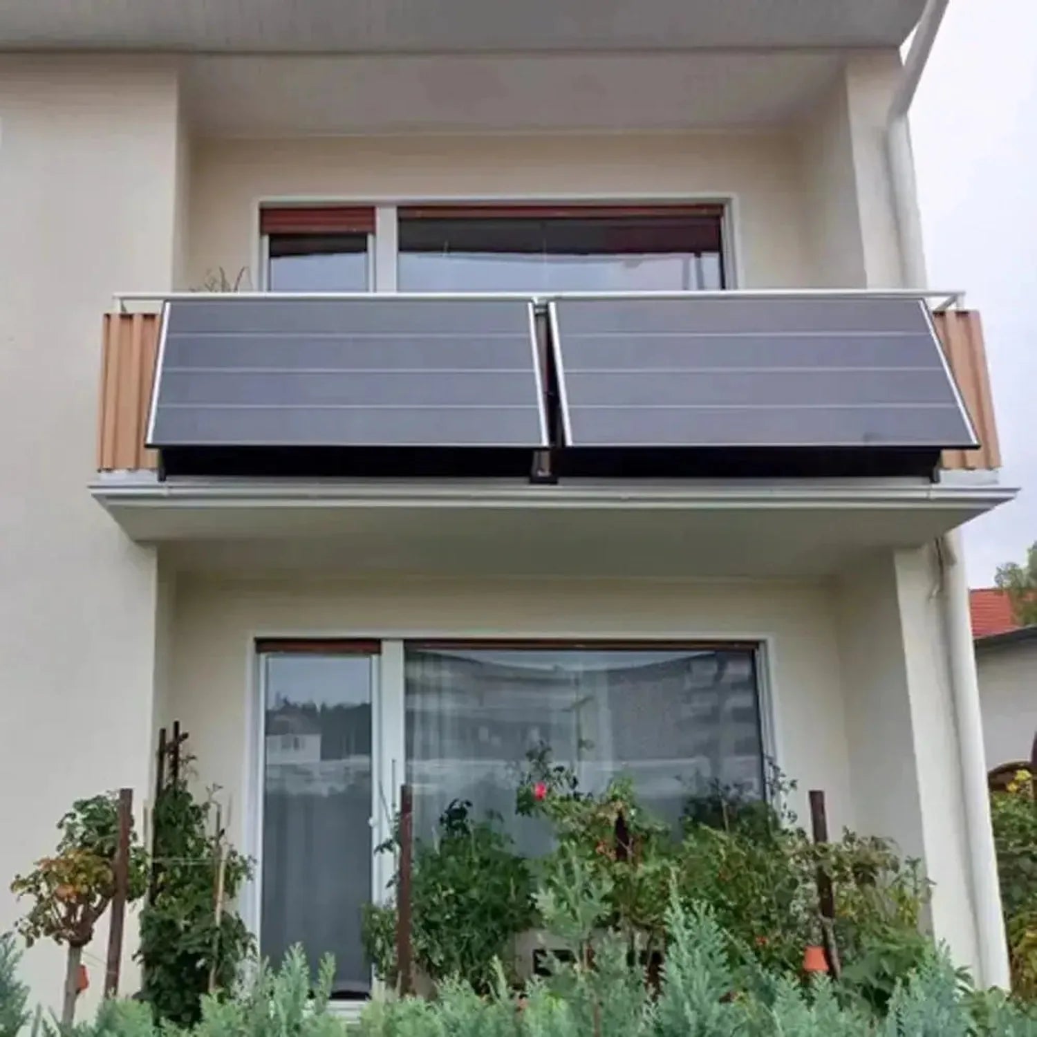Two solar panels mounted on a modern white building's balcony railing, angled toward the sun with adjustment brackets visible