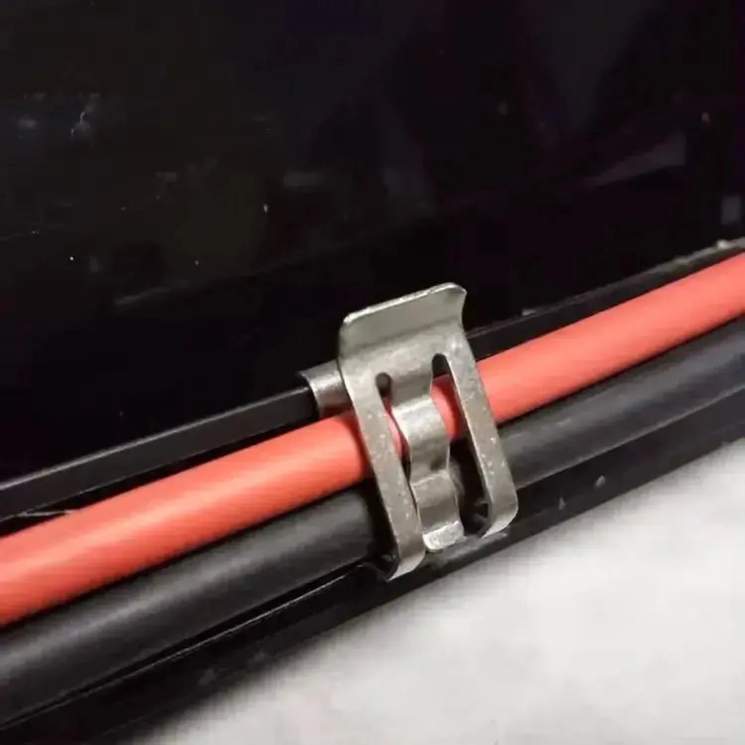 Stainless steel cable clip securing red and black wires to black frame rail, installation shot.