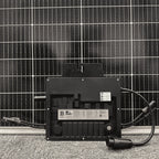 Black microinverter with cable connectors mounted on gravel in front of solar panel array, showing specifications label and Q