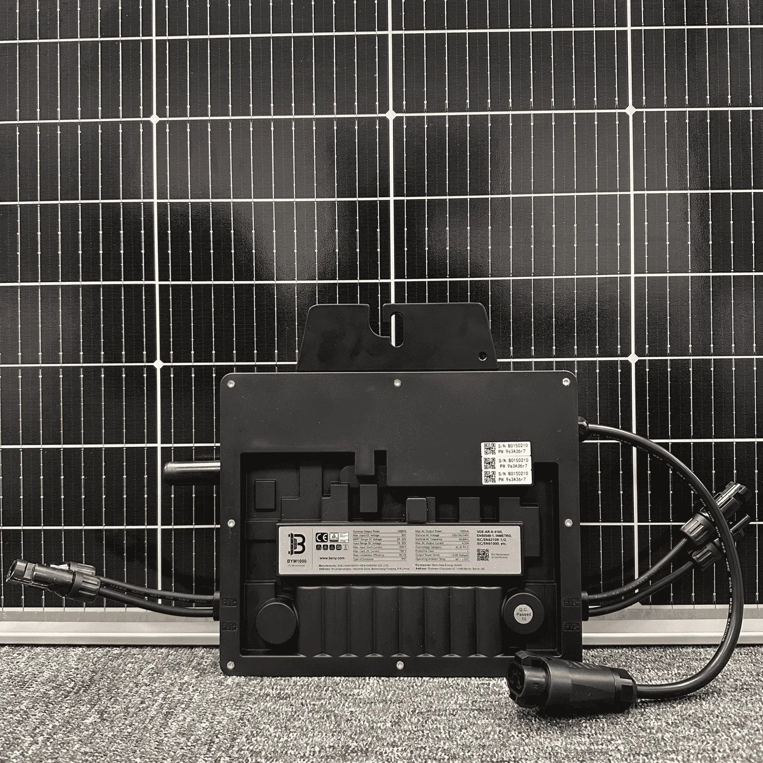 Black microinverter with cable connectors mounted on gravel in front of solar panel array, showing specifications label and Q