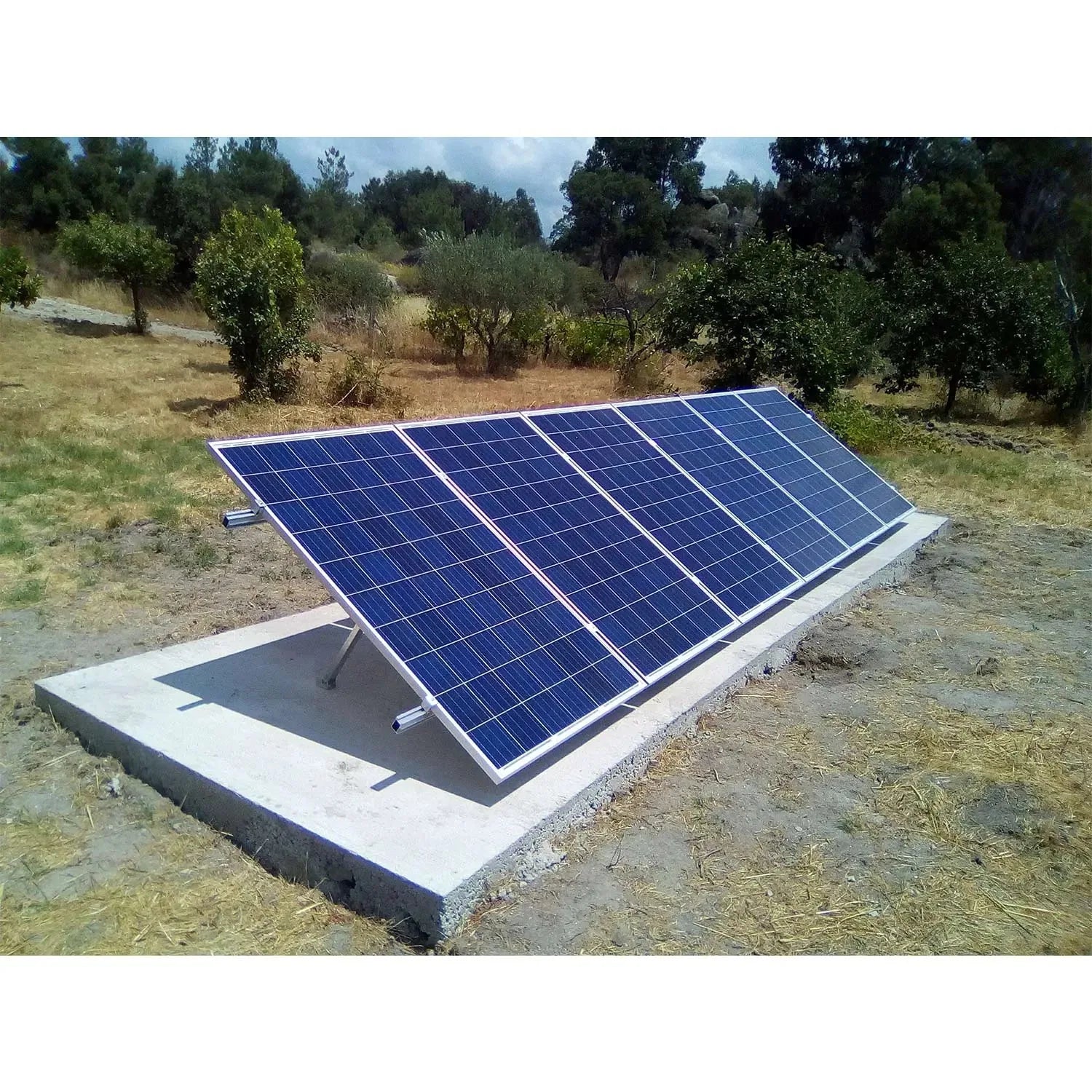 Five solar panels mounted on adjustable metal frame on concrete foundation in rural landscape with trees.