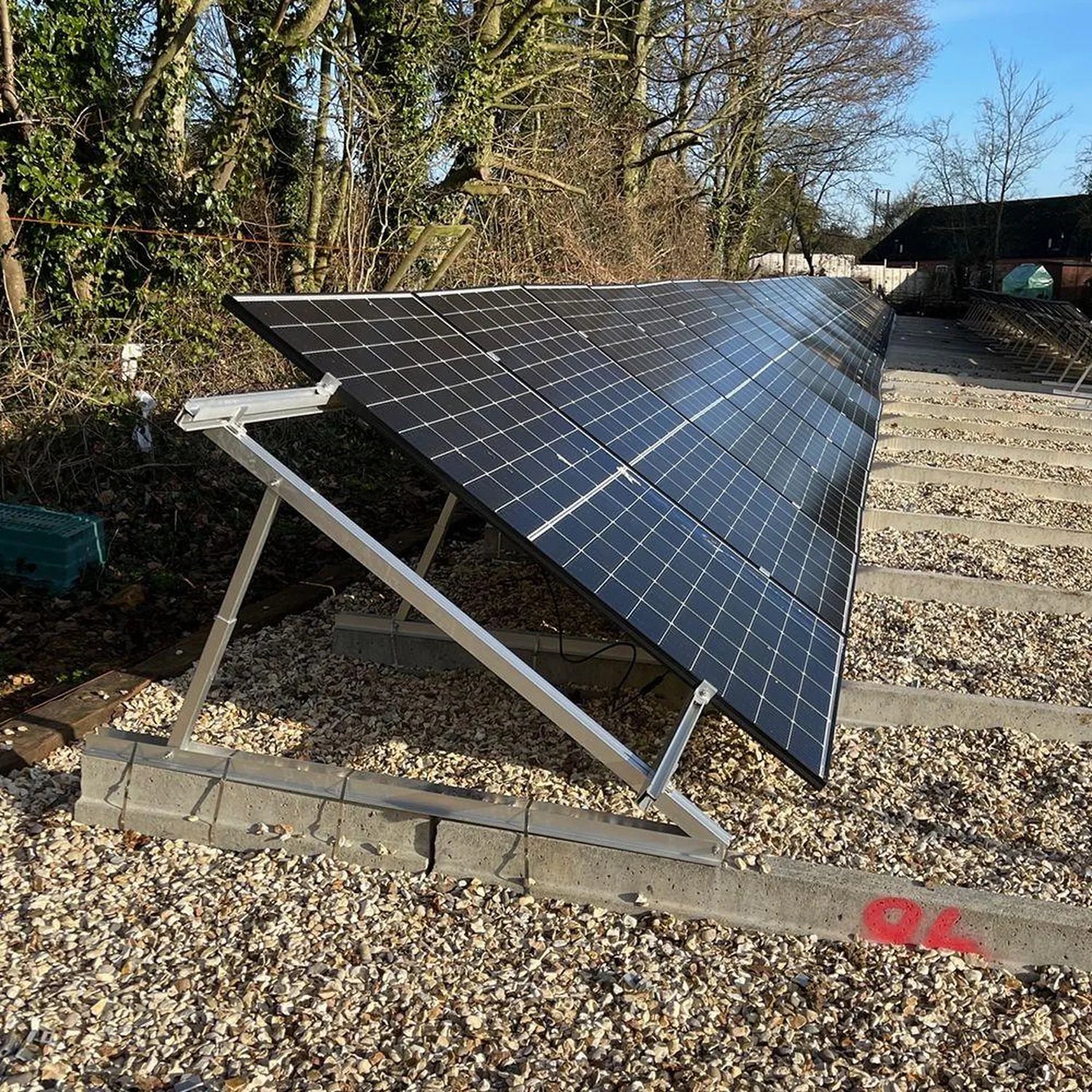 Adjustable solar panel mounting kit installed on gravel ground with panels angled toward sunlight, trees and farm buildings v