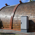 Metallic battery cabinet installed outdoors beside modern wooden architecture with curved metal detailing.