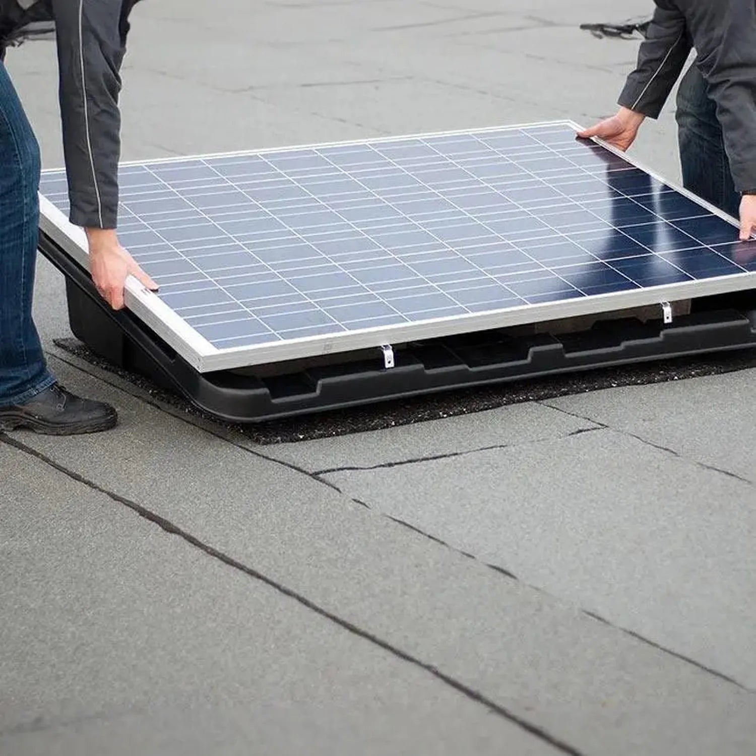 Two people installing a solar panel on a flat roof using a black mounting tray system with white frame brackets.