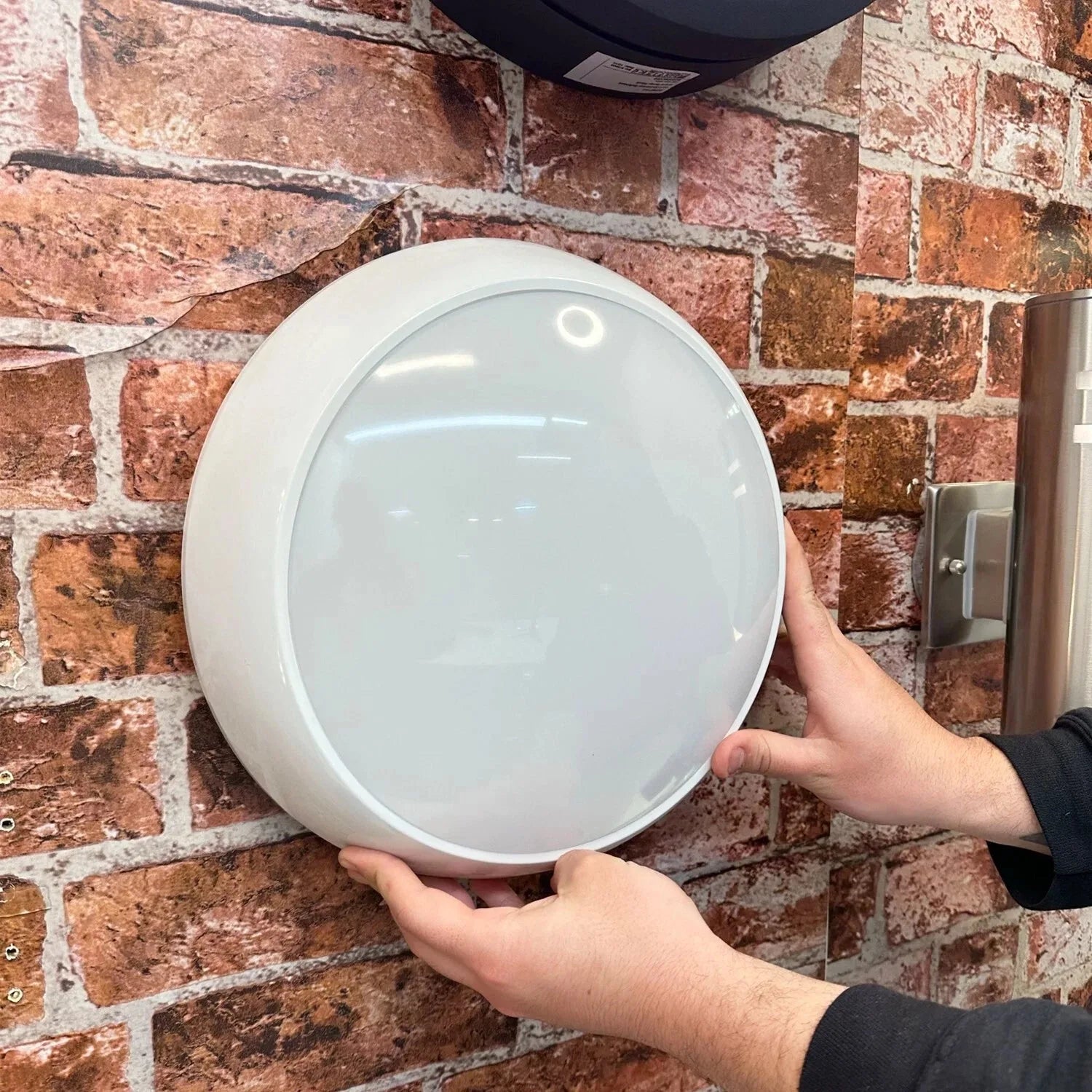 Person holding white circular LED retrofit lamp against red brick wall installation background.
