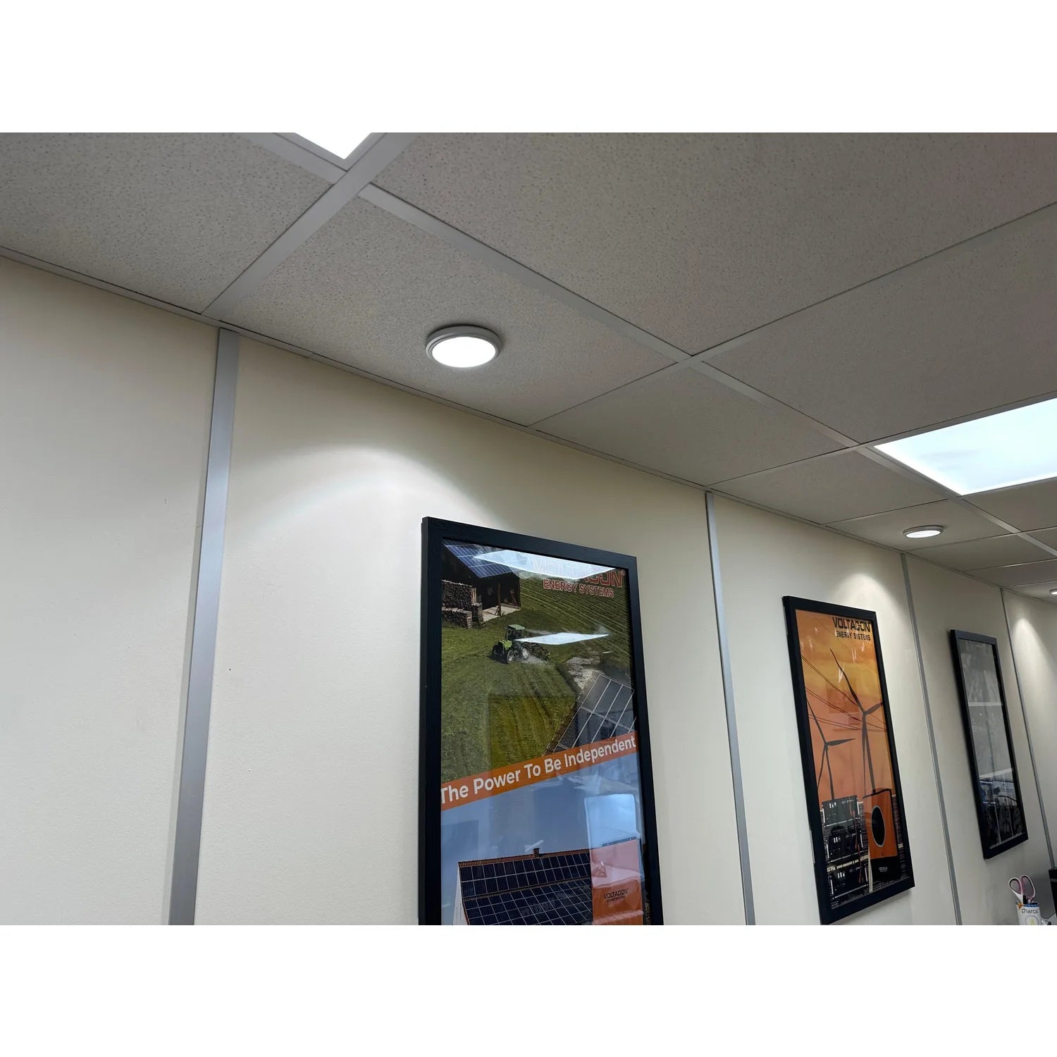 Recessed LED ceiling light installed in office hallway with drop ceiling tiles, illuminating wall-mounted energy posters belo