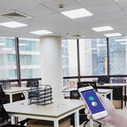 Hand holding smartphone with smart control app in modern office with LED panel ceiling lights and city views