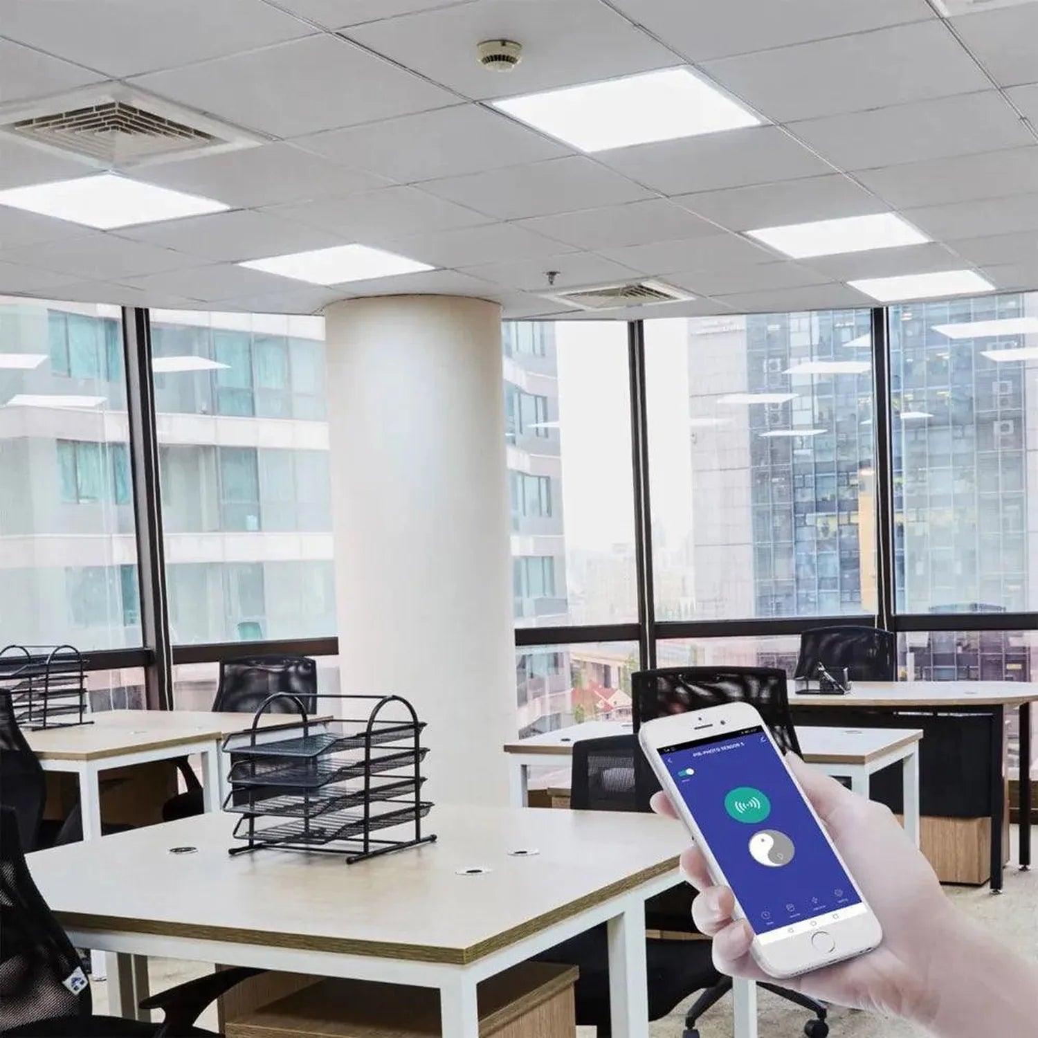Hand holding smartphone with smart control app in modern office with LED panel ceiling lights and city views