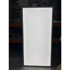 Rectangular LED panel light with white illuminated face and dark frame, displayed vertically indoors against shelving.