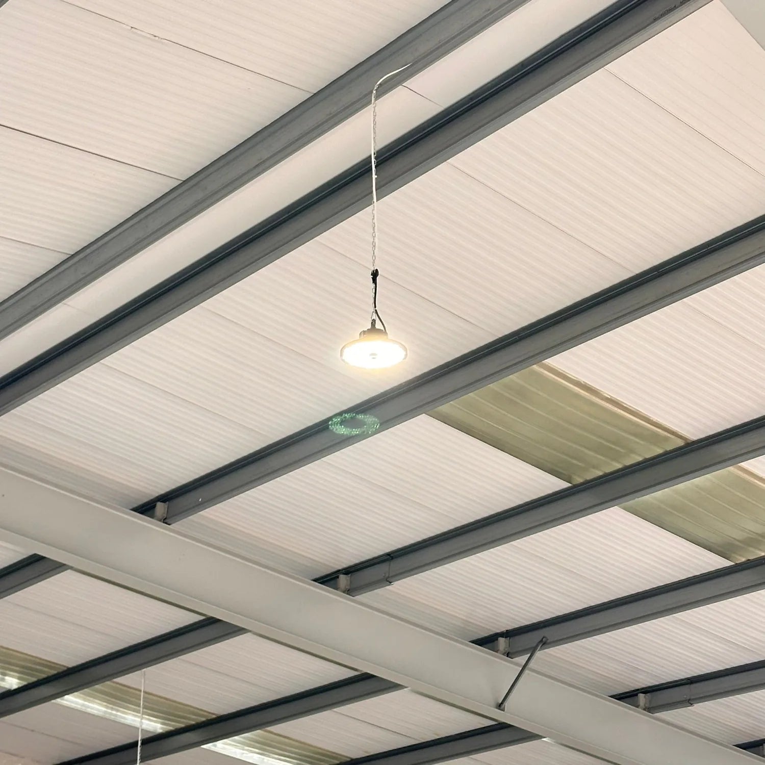LED high bay light suspended from industrial ceiling beams in warehouse installation shot.