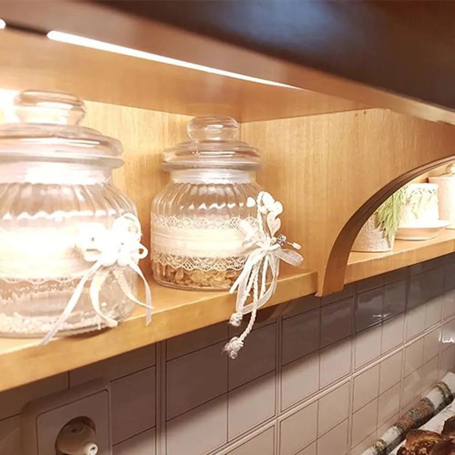 Glass storage jars with lace trim displayed on wooden shelf under warm LED lighting in kitchen cabinet installation.