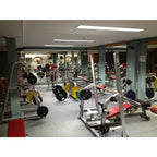Interior gymnasium with weight training equipment, barbells, and exercise machines under fluorescent ceiling lighting.