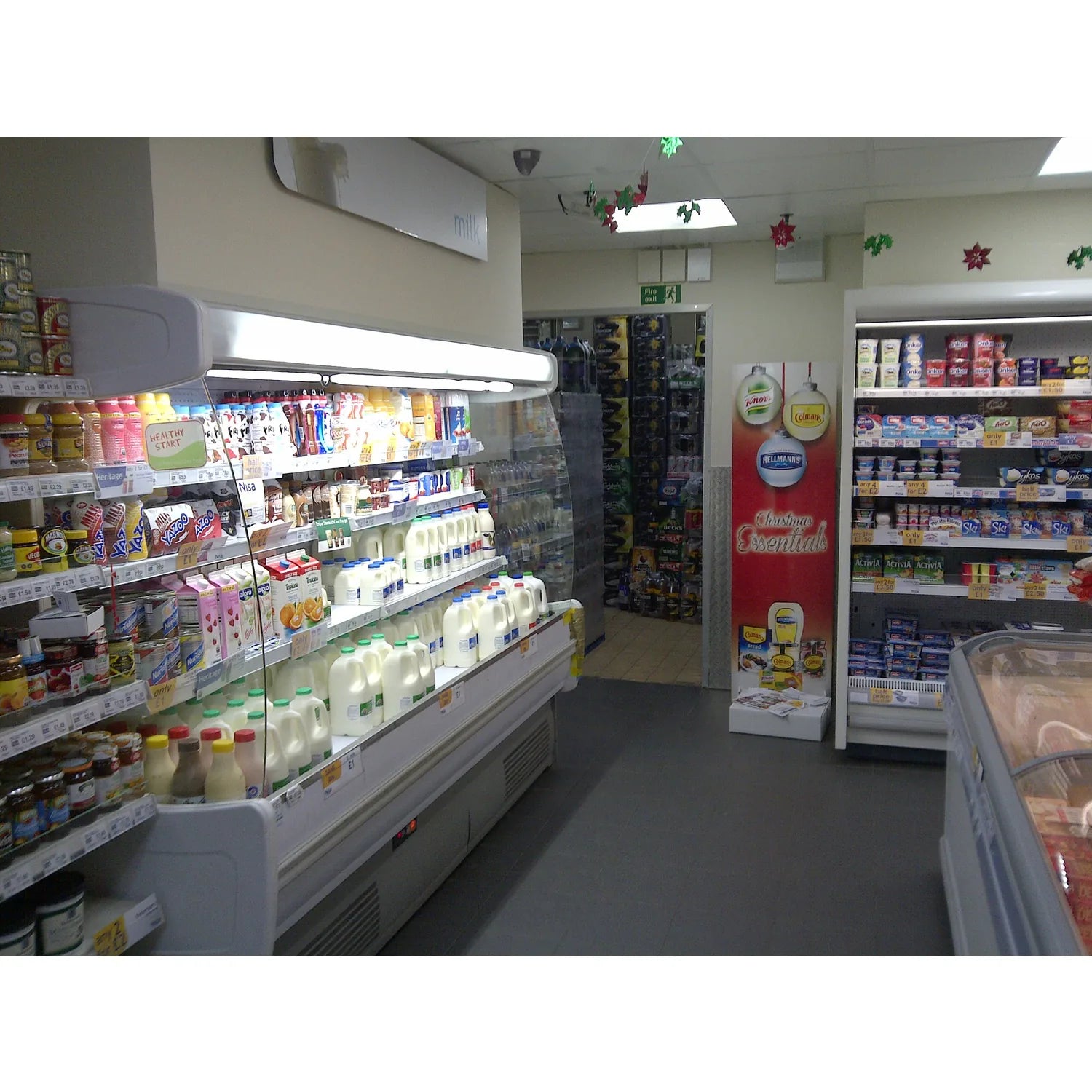 Refrigerated dairy display case in convenience store, lit by fluorescent overhead lighting and bright LED tube lights along t