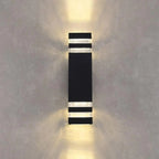 Black cylindrical LED wall light mounted on white wall, glowing with warm white light radiating upward and downward.