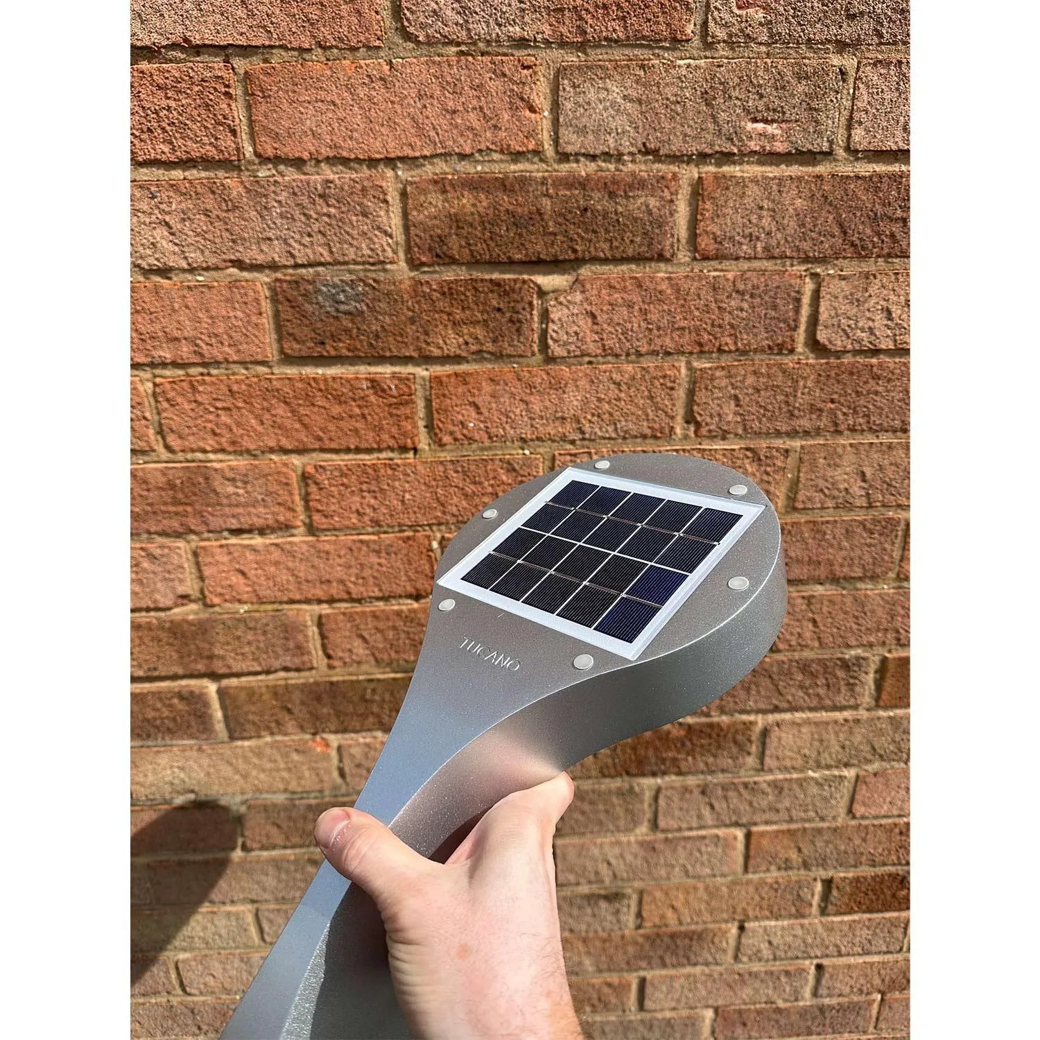 Hand holding gray solar wall light with small solar panel against brick wall background.