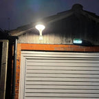 Solar LED wall light mounted above garage door at dusk, illuminating orange brick and metal corrugated building with warm whi