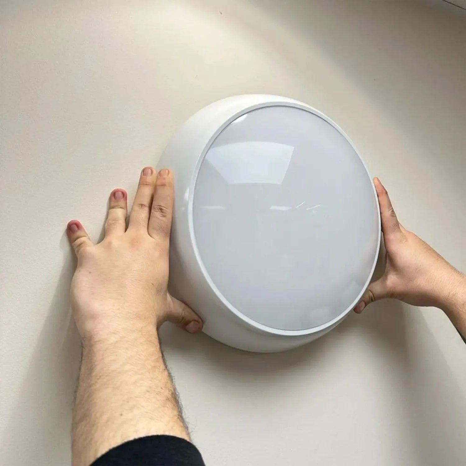 Person holding oval white LED bulkhead light fixture against beige wall, showing front face with frosted diffuser panel.
