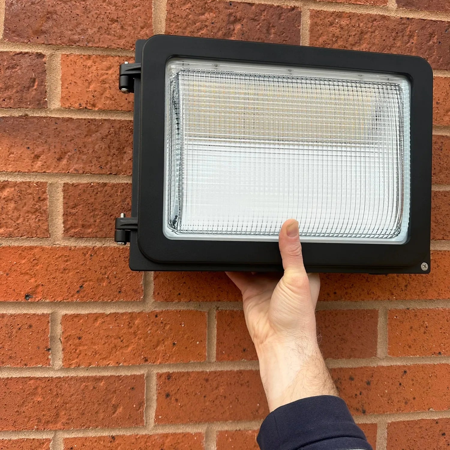 Person holding black rectangular LED wall pack light mounted on red brick wall, installation shot.
