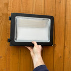 Hand holding rectangular black LED wall light with clear lens against wooden wall, showing installation setup