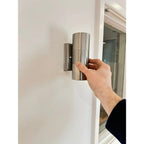 Hand holding brushed stainless steel cylindrical outdoor wall light mounted on white wall near glass door.