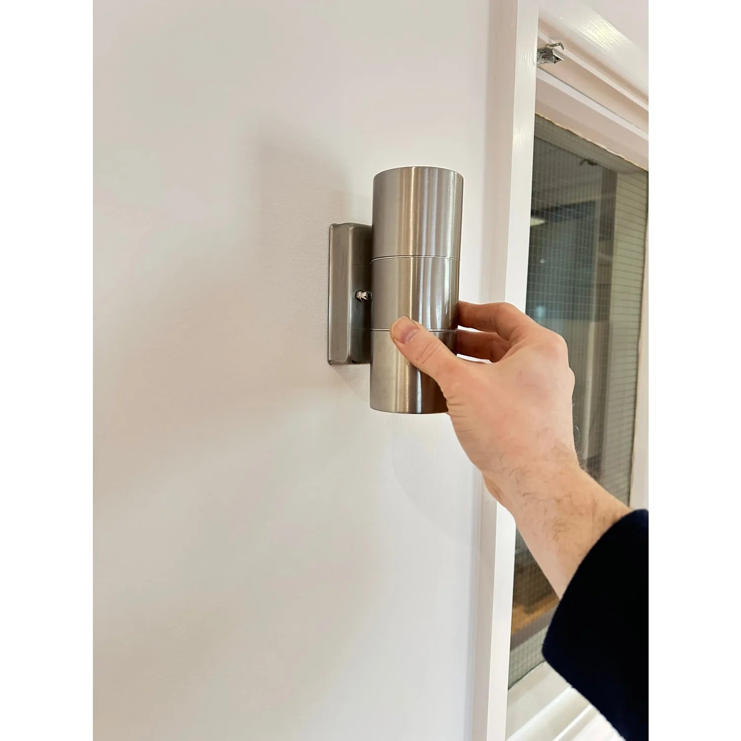 Hand holding brushed stainless steel cylindrical outdoor wall light mounted on white wall near glass door.