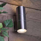 Cylindrical stainless steel outdoor wall light mounted on brick wall with warm light glowing from bottom opening, ivy visible