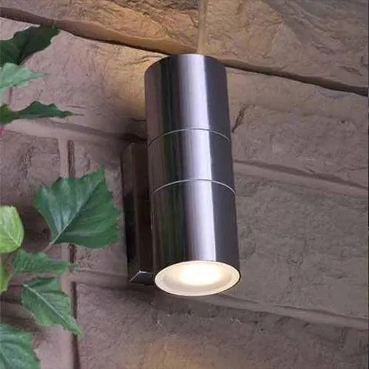 Cylindrical stainless steel outdoor wall light mounted on brick wall with warm light glowing from bottom opening, ivy visible