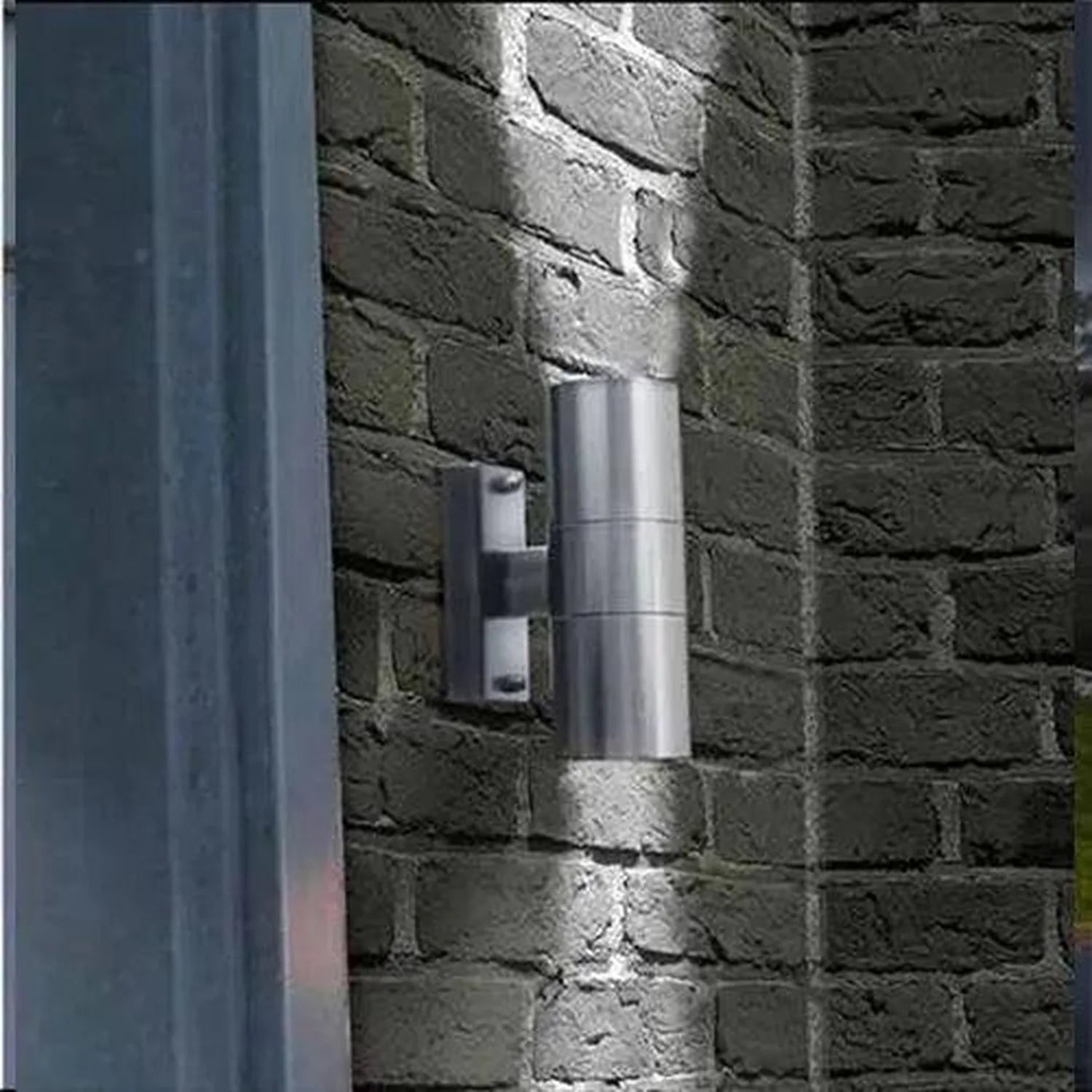 Stainless steel cylindrical wall light mounted on dark brick wall, showing twin pack installation shot with lighting effect.