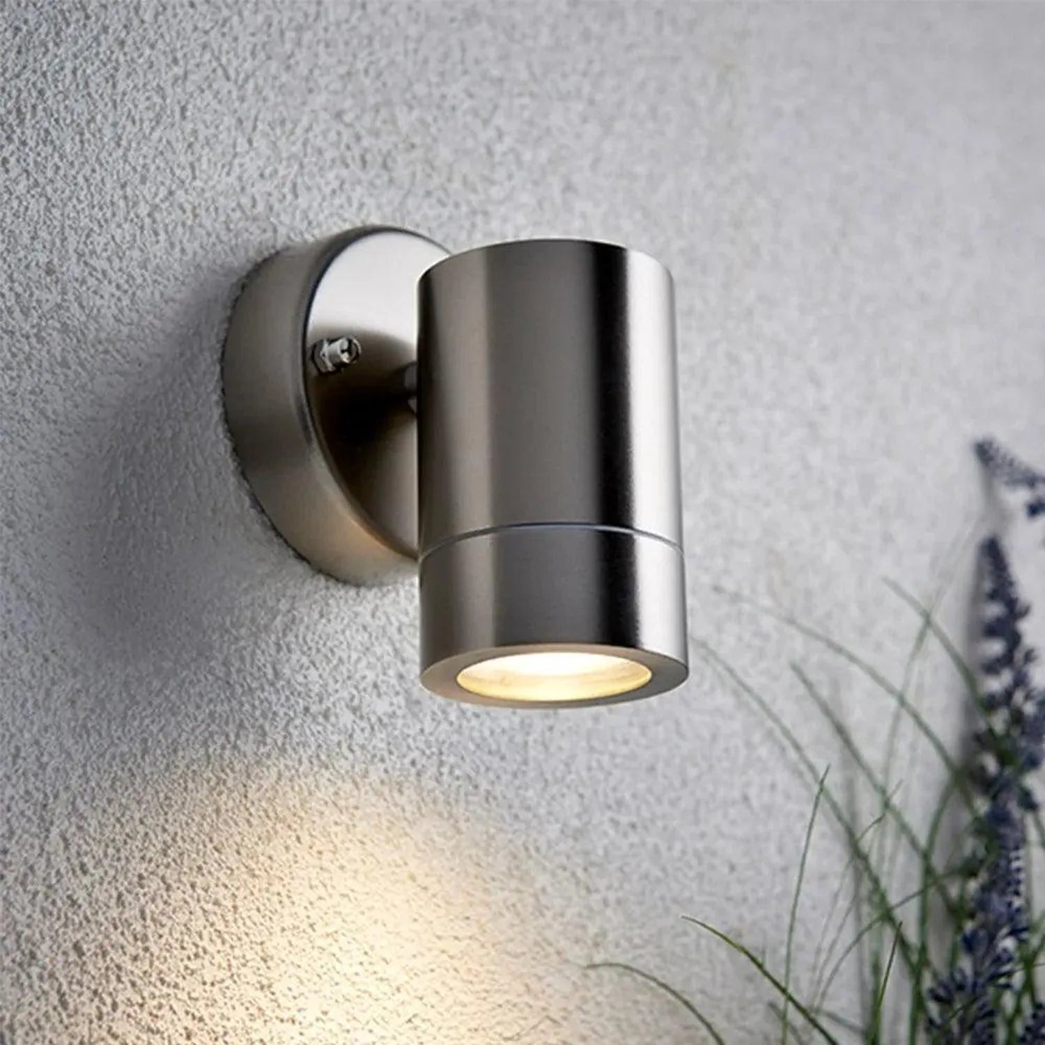 Stainless steel cylindrical downlight mounted on textured wall, illuminating ground below, with purple flowers visible at edg