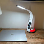 White curved LED desk lamp with digital clock display and red charging base, illuminating a workspace with laptop.