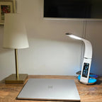 OttLite Wellness LED desk lamp with white curved shade and digital clock display on wooden desk next to laptop and smartphone