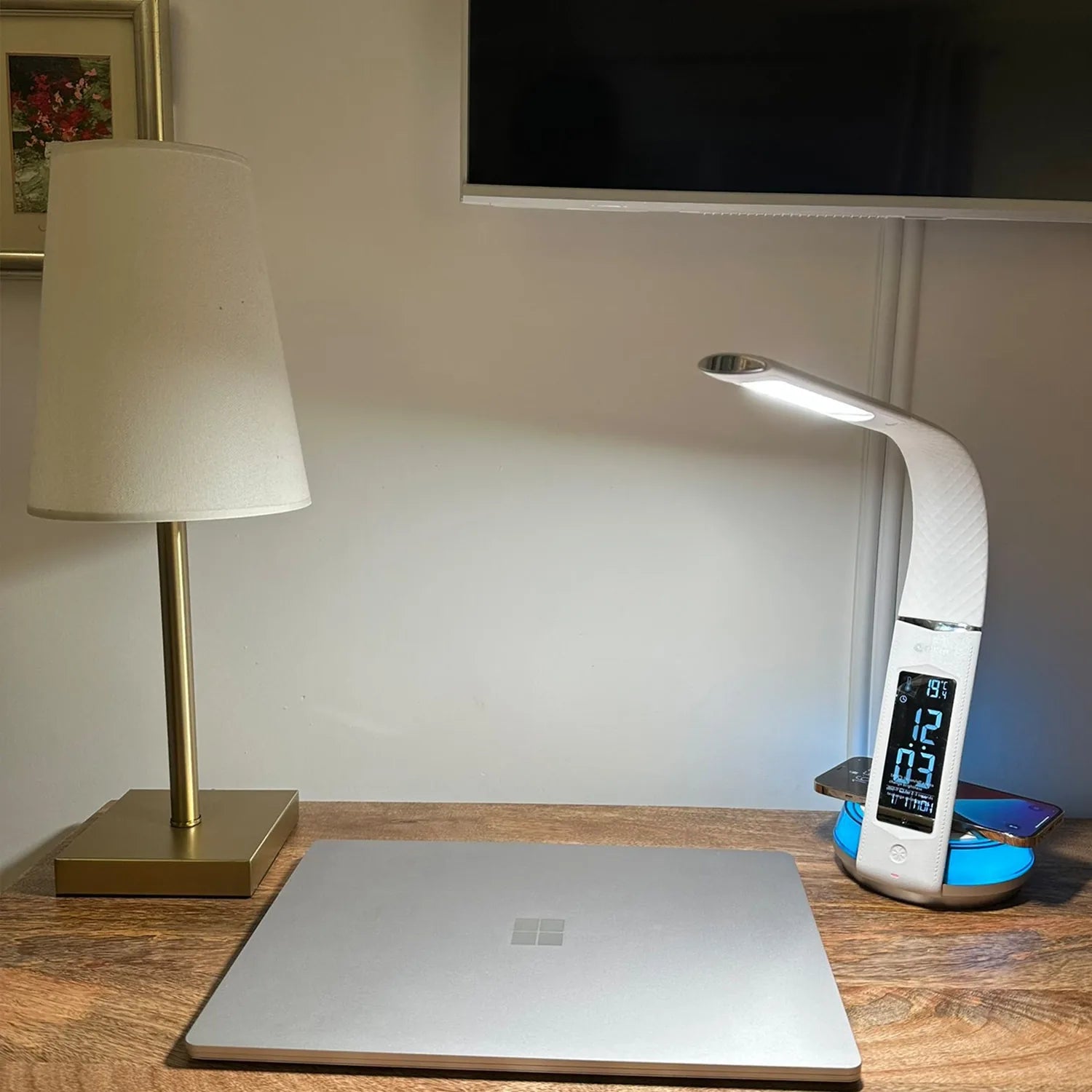 OttLite Wellness LED desk lamp with white curved shade and digital clock display on wooden desk next to laptop and smartphone