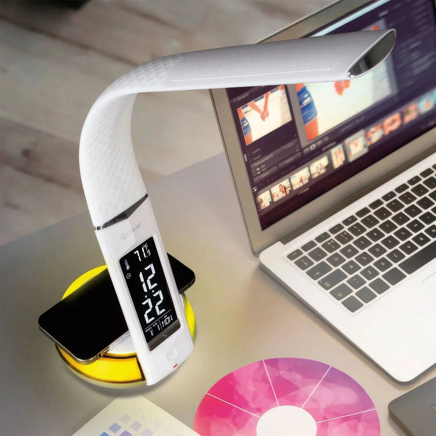 White LED desk lamp with digital clock display on yellow charging base, positioned next to laptop and wireless charging pad o