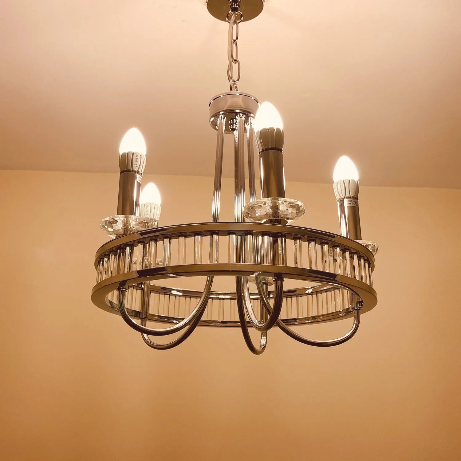 Modern brass chandelier with five lit LED candle bulbs and crystal accents, hanging from chain against beige ceiling.