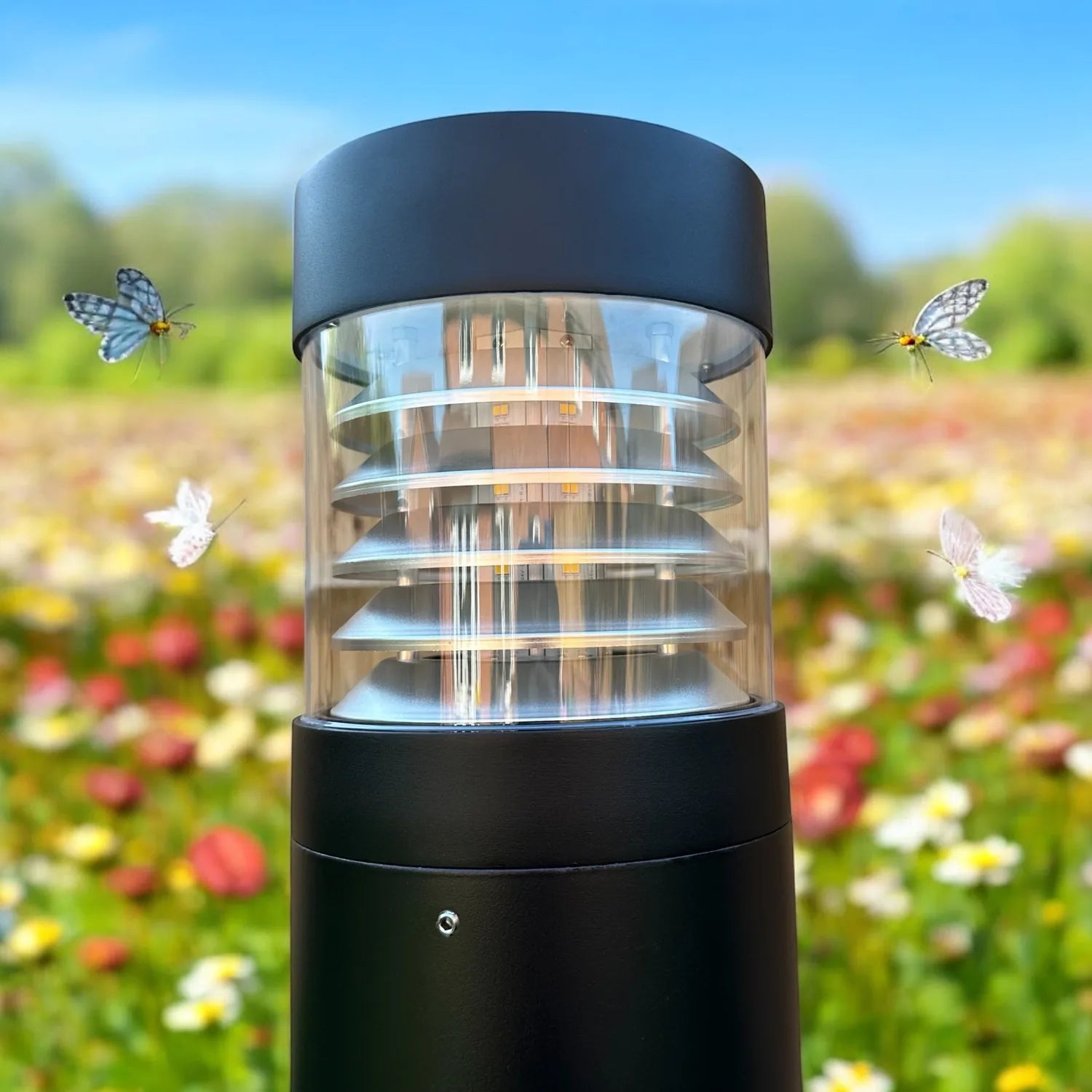 Black cylindrical LED bollard light with louvered cylindrical diffuser, shown installed in flowering garden with butterflies 