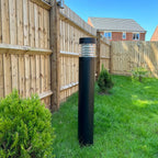 Black LED bollard light installed in residential garden next to wooden fence and brick house.