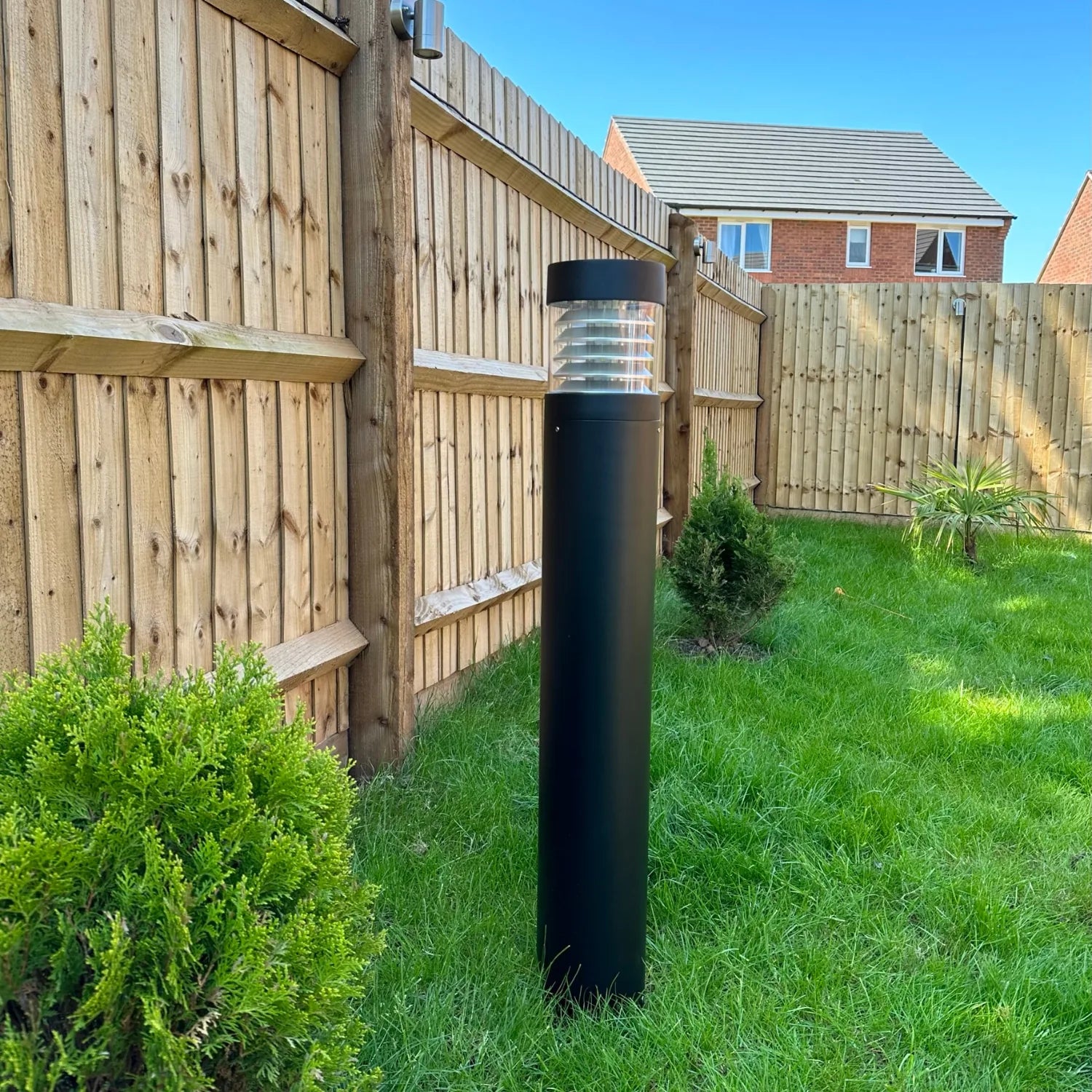 Black LED bollard light installed in residential garden next to wooden fence and brick house.