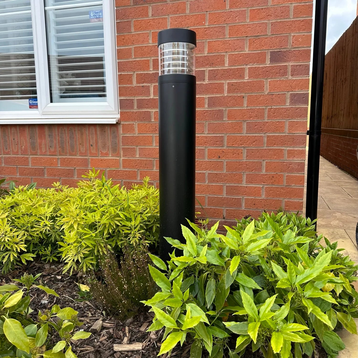 Black cylindrical LED bollard light installed in garden bed surrounded by green shrubs against red brick wall.