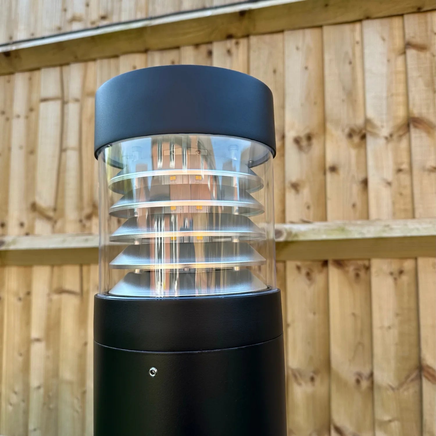 Black cylindrical LED bollard light with clear polycarbonate louvered diffuser, mounted against wooden fence background, show