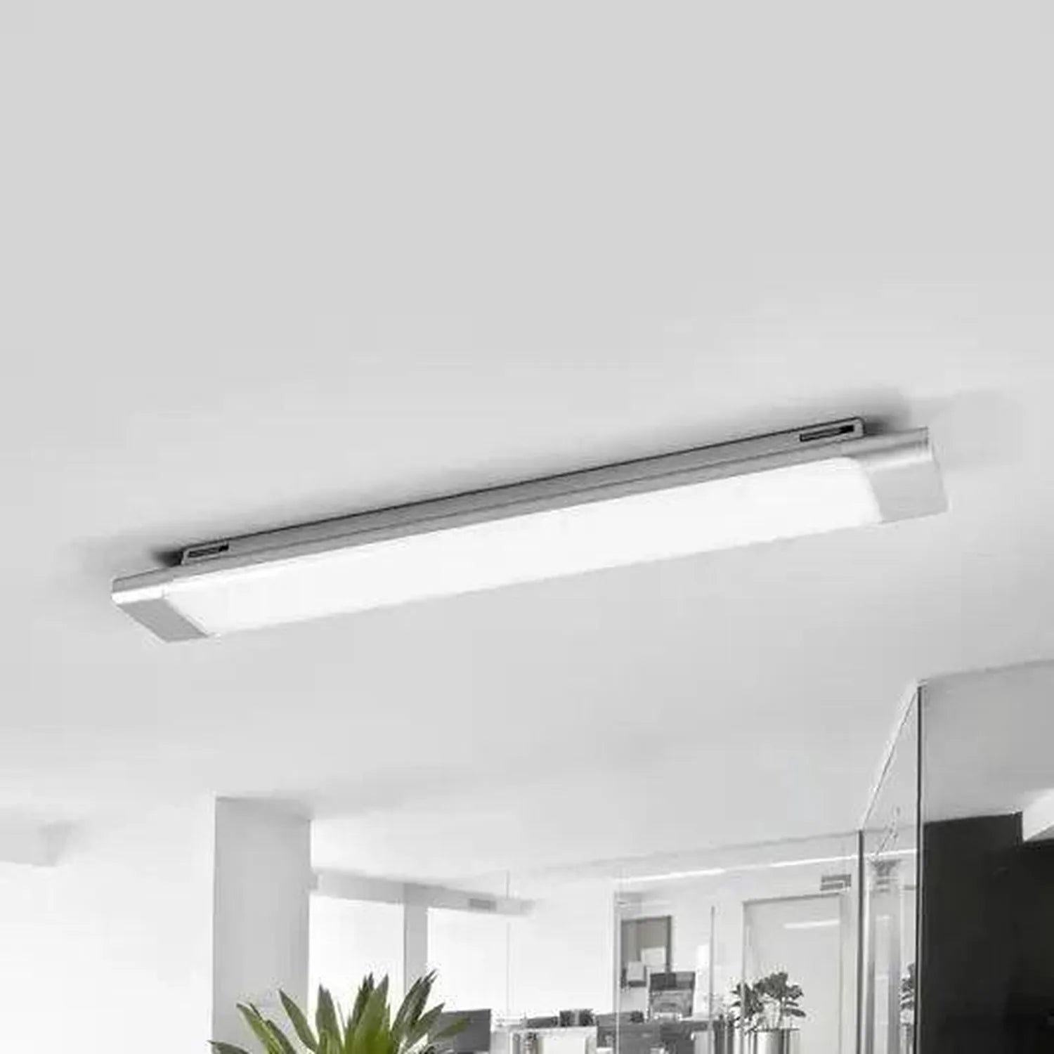 Sleek grey aluminium LED batten light mounted horizontally on white ceiling in modern office space with plants and glass part