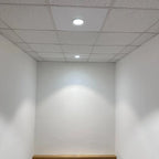 Recessed LED downlight installed in drop ceiling tile, illuminating white walls and wooden floor in empty room.