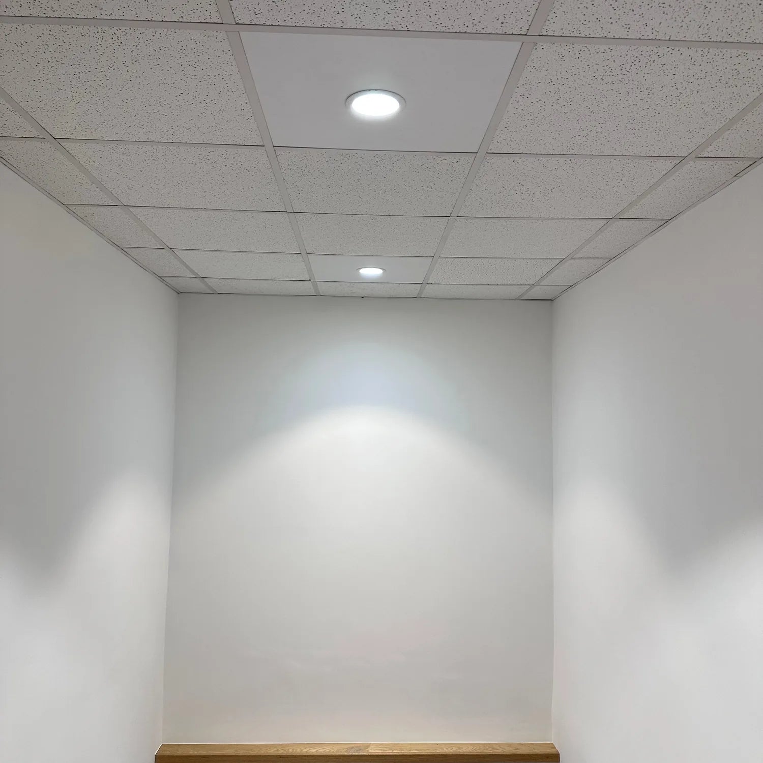 Recessed LED downlight installed in drop ceiling tile, illuminating white walls and wooden floor in empty room.